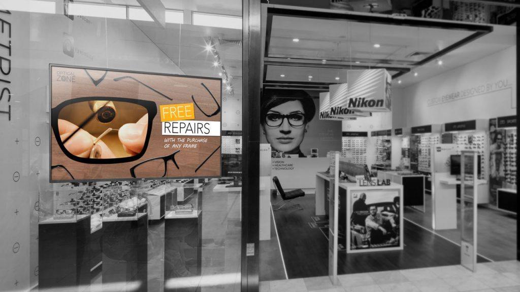 Digital signage TV display mounted inside the front window of an eyewear store showing a promotion for free repairs