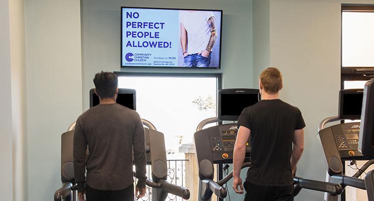 Two men on treadmills with a digital signage tv above them promoting services